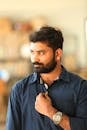 A thoughtful man with a beard and blue shirt posing indoors, displaying a stylish watch.